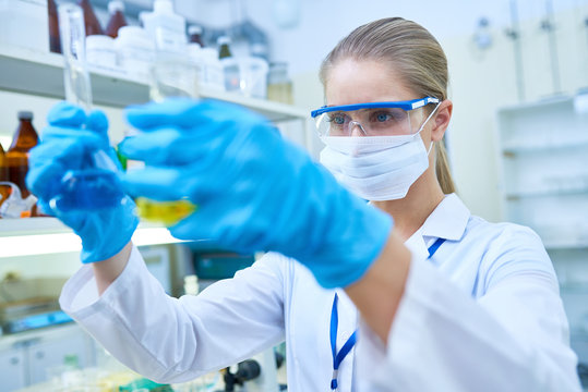 Serious thoughtful young female scientist wearing surgical mask and protective goggles analyzing color of liquids in flasks while working in laboratory