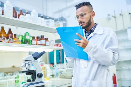 Serious confident young Arabian scientist with beard wearing eyeglasses making notes in folder while keeping records about scientific experiment in laboratory