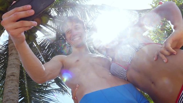 Romantic happy couple in the sunlight taking selfie on cell smart phone hugging outdoors under palm trees on the sun and sun slare on the background. slwo motion. 1920x1080
