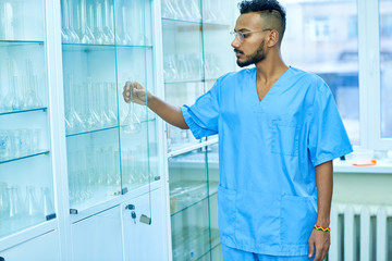 Serious young Arabian technician with beard wearing uniform taking flask from cupboard in laboratory