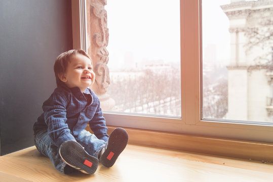 Little happy kid in casual look sitting and smile indoors. Baby boy sit at window sill with sunlight.