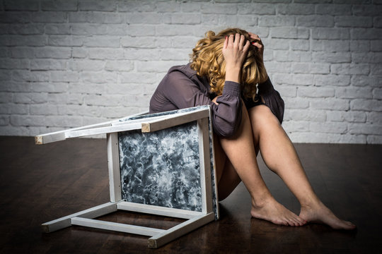  The wooden chair is tilted and lies on the floor. Woman sitting beside the chair. The woman is wearing a long shirt.
