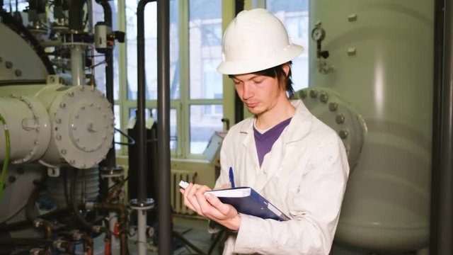 The inspector in a white helmet and a robe examines the gas installations in the laboratory, makes notes in the magazine.