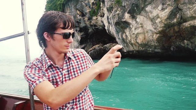 Young handsome man in sunglasses takes photo with phone in boat travel against rocky island in azure bay. slow motion. 1920x1080