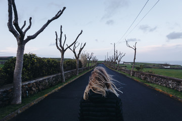 Young woman standing on a black road in countryside lined with stone wall and trees without leaves...