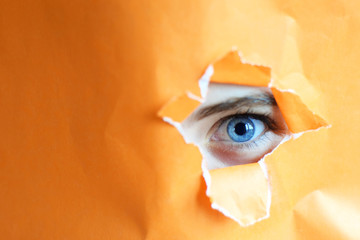 Teen girl's blue eye looking through hole in orange paper.