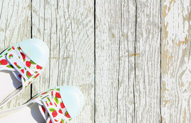 Obraz premium White with red berries sneakers on a white wooden background. Summer shoes top view. Colored slippers stand on old painted boards. Free space for text.