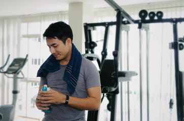 muscular asian young handsome man wipes sweat with towel and drinking water while resting after workout for good healthy in fitness gym at morning, bodybuilder, lifestyle and sport exercise concept