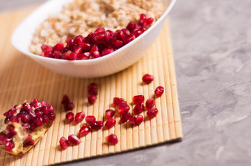 Cooked yummy warm oatmeal and heap of ripe fresh pomegranate seeds in white ceramic bowl near piece of pomegranate on clean bamboo rug on worn gray scratched concrete with copy space