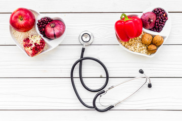Products good for heart and blood vessels. Vegetables, fruits, nuts in heart shaped bowl near stethoscope on white wooden background top view copy space