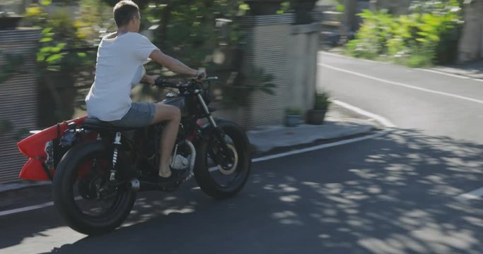 Handsome man biker surfer driving his black motorcycle cafe racer with red surfboard shortboard on road at sunny day. 4k video shooting by handheld gimbal