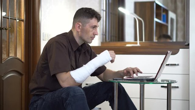 A man with a broken right hand in a plaster sits near a coffee table with a laptop in a room with furniture. Broken arm