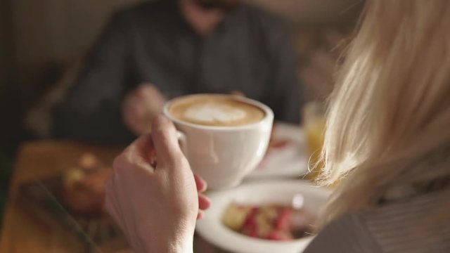 Close up shot of blonde woman drinking a cup of coffee in a cafe with her boyfriend. Blured dishes on the table.