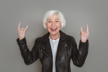 Senior woman in leather jacket studio isolated on grey wall showing horns sign
