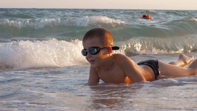 Slow motion shot of a boy in goggles lying on the shore and enjoying sea waves rolling in and washing him, scene at sunset. Summer vacation at the seaside
