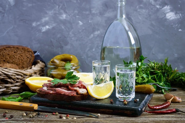 Two shot glasses of vodka with lemon slice, pickled cucumbers and rye bread with salted bacon on the dark background. Traditional strong drink and appetizer. Close-up.