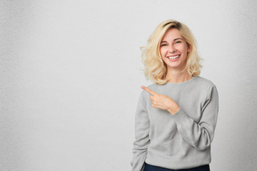 Happy excited female with blonde hair indicates with fore finger aside, demonstrates something at...