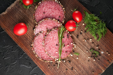Smoked salami on a black table and a brown board. Sausages with rosemary, garlic and pepper and tomato cherry, dill, parsley.