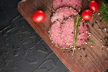 Smoked salami on a black table and a brown board. Sausages with rosemary, garlic and pepper and tomato cherry, dill, parsley.