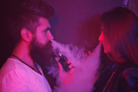 Man and woman standing smoking e-cigarettes with lowe angle view off the middle-aged man exhaling smoke from his nostrils in the foreground.They rest in vapeshop. They let the smoke out of his mouth