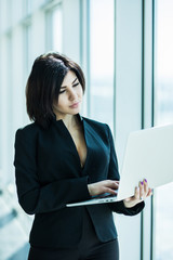 Businesswoman standing against office window holding laptop. Businesswoman in bright office.