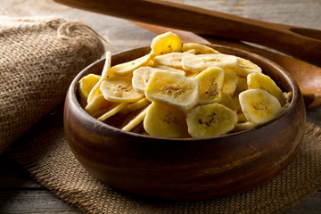 Heap of dried banana chips snack in wooden bowl on dark table