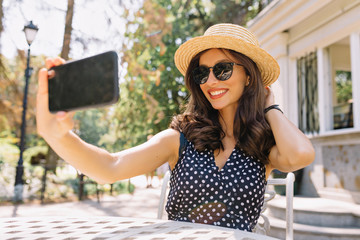 Pretty lovely young woman dressed hat and black sunglasses makes a selfie on smartphone in the park in summer weather 