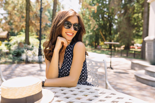 Image of young woman with beautiful hair and charming smile is sitting in summer cafeteria in sunlight. She is wearing pretty summer dress and black sunglasses. Background park. Beautiful portrait.
