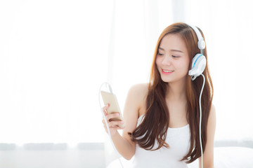 Beautiful asian young woman enjoy listen music with headphone and holding smart mobile phone while sitting in bedroom, relax girl with earphone, leisure and technology concept.