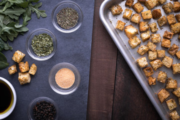 Top down view of ingredient dishes of spices on slate, beside a sheet pan of croutons