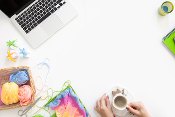 The girl knits the colored canvas with knitting needles. Laptop, basket with skeins of yarn, coffee, tea and water with lemon.