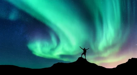 Poster Aurora and silhouette of standing man with raised up arms on the mountain in Norway. Aurora borealis and happy man. Starry sky, green polar lights. Night landscape. Northern lights. Travel and tourism © den-belitsky