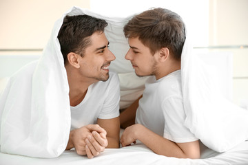 Young gay couple lying under blanket on bed in light room