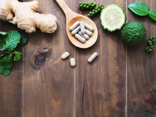 herbal pills in wooden spoon with ginger root, Kaffir lime fruit and flower on dark brown wood background with copy space. Above view.