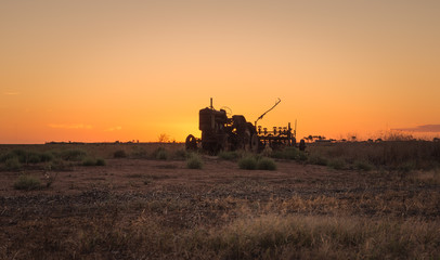 Fototapeta premium Old abandoned tractor in farm field with rusted agricultural equipment silhouetting at sunset