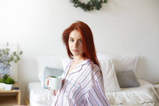 Picture of attractive sleepy young red haired European female wearing stylish striped night suit, sitting in her cozy bedroom, drinking fresh strong coffee early in the morning before work