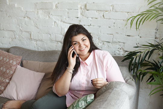 People, modern technologies and communication concept. Picture of beautiful overweight happy young brunette woman wearing pink shirt smiling happily while talking on mobile phone with her boyfriend