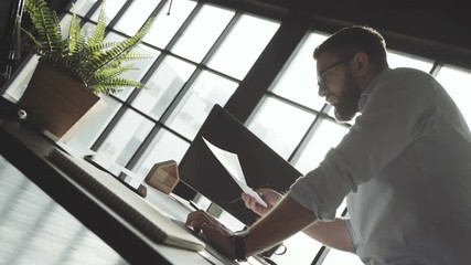 Man with a beard in white shirt works against the background of a large panoramic window, leans on table. Young businessman works alone in bright spacious office, stylish accessories and plants.