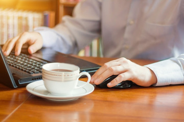 Young caucasian businessman working in office. Using internet in laptop. Cup of coffee on the table. Library books on background.