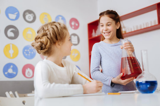 Pleasant conversation. Charming teenage girl talking to her friend during the chemistry class while holding a big flask with a red liquid