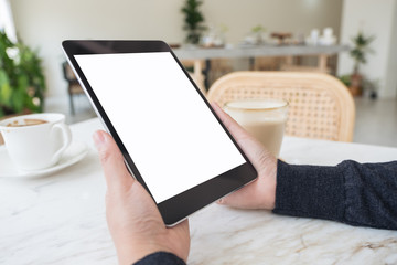 Mockup image of hands holding black tablet pc with blank white screen and coffee cup on table in cafe