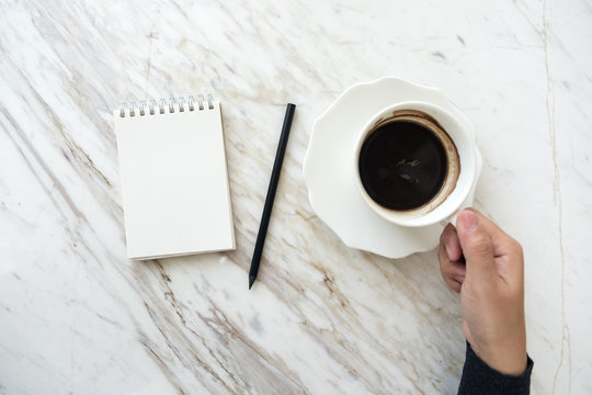 Top view image of a hand holding coffee cup with a blank white notebook on table
