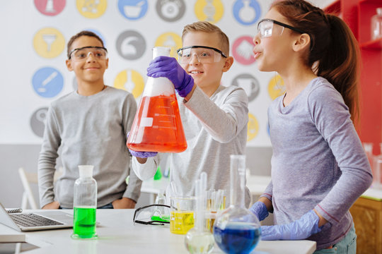 Check it out. Upbeat teenage boy lifting up a big flask and showing the ongoing reaction inside it to his classmates while they having a chemistry class in the lab together