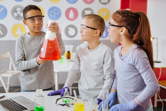 Exciting experiment. Charming teenage boy holding a big flask and demonstrating the chemical reaction to his classmates while they all wearing safety goggles