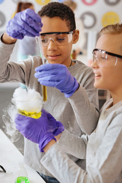Best cooperation. Cheerful teenage boys working on a chemical experiment together during class while one of the boys holding a fuming flask and the other adding chemical to test tube