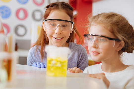 Fascinating chemical reaction. Cheerful teenage female students squatting near the table and watching the smoke coming out of a chemical flask with awed expressions