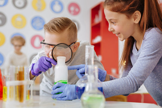Examining thoroughly. Joyful teenage boy examining a chemical substance in the flask with a magnifying glass while his classmate holding that flask for him