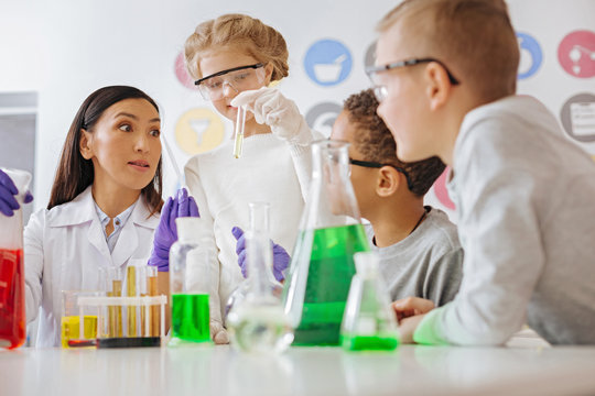 Little prodigies. Pleasant young chemistry teacher holding at a pipette with a chemical and looking at test tube in her students hands while looking surprised by her level of knowledge