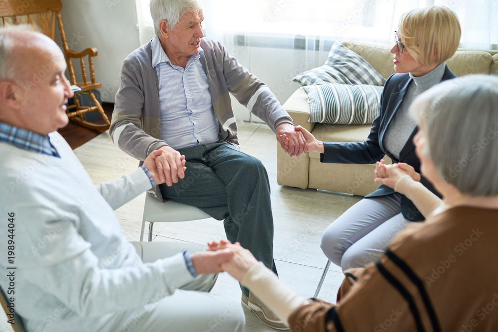 Wall mural Portrait of senior people holding hands in group therapy session lead by female psychiatrist, copy space