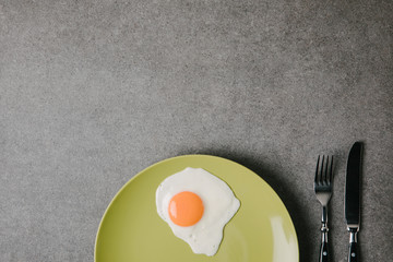 top view of fresh fried egg on plate and fork with knife on grey